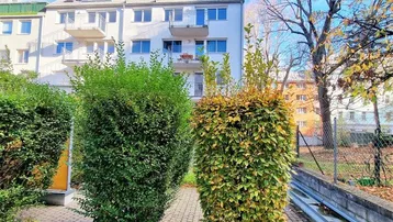 Weißes Mehrfamilienhaus mit Balkonen, umgeben von gepflegten Hecken und Bäumen unter blauem Himmel.