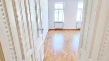 Heller Wohnraum mit Parkettboden und hohen Fenstern, gesehen durch eine offene Tür.