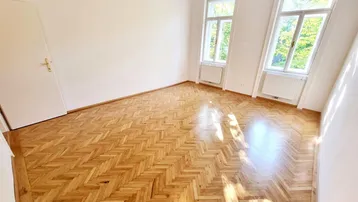 Helles Zimmer mit Fischgrätparkettboden, weißen Wänden und zwei Fenstern, die viel Tageslicht hereinlassen.
