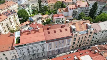 Panoramablick über eine dicht bebaute Stadtlandschaft mit zahlreichen Gebäuden und roten Dächern, umgeben von Grünflächen.