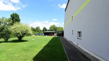 Seitenansicht des Gebäudes mit großem Rasenbereich, Bäumen und einem Carport.
