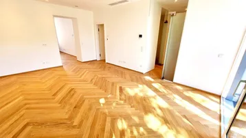 Heller, leerer Raum mit Fischgrätparkett und großen Fenstern, die viel Tageslicht hereinlassen.