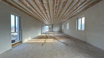 Großer, unfertiger Raum mit sichtbaren Deckenbalken, Rohbetonboden und viel Tageslicht durch mehrere Fenster.