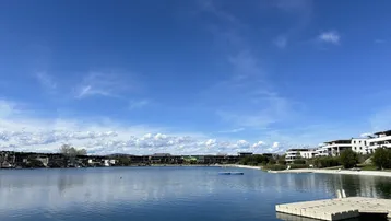 Blauer Himmel über einem See mit modernen Gebäuden am Ufer und einem Steg im Vordergrund.