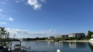 Weiter Blick über einen ruhigen See mit modernen Wohngebäuden im Hintergrund unter blauem Himmel.
