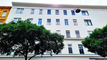 Weißes Mehrfamilienhaus mit vielen Fenstern und Bäumen davor unter bewölktem Himmel.