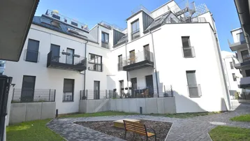 Moderner Apartmentkomplex mit weißer Fassade, Balkonen und einem gemeinschaftlichen Gartenbereich mit Sitzbank.