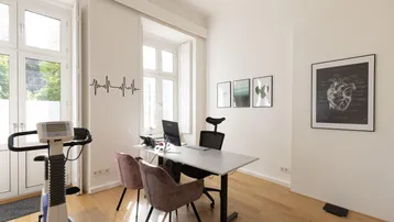 Helles Büro oder Behandlungszimmer mit großem Fenster, Schreibtisch und Trainingsgerät.