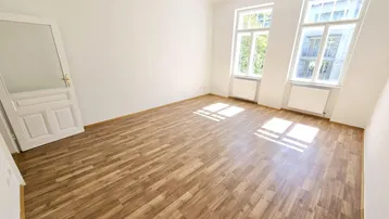 Helles Zimmer mit Laminatboden und zwei großen Fenstern, das viel Tageslicht hereinlässt.