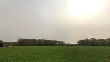 Weitläufiges grünes Feld unter bewölktem Himmel mit Baumreihe im Hintergrund, ideal für ländliche Ruhe.