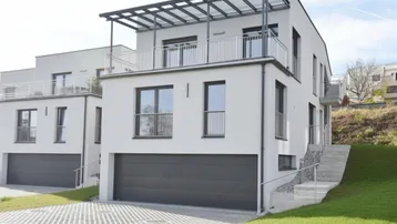 Seitenansicht eines modernen Neubaus mit Garage, Balkon und gepflegter Grünfläche am Hang.