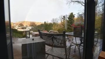 Blick von innen auf eine Holzterrasse mit Gartenmöbeln und einem Regenbogen am Horizont.