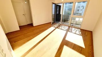Helles, leeres Zimmer mit Holzboden und Zugang zu einem Balkon, durchflutet von Sonnenlicht.
