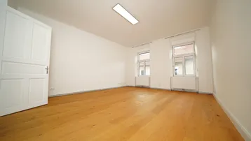 Helles, leeres Zimmer mit Parkettboden und zwei Fenstern, bereit zur individuellen Gestaltung.