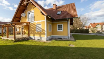 Gelbes Einfamilienhaus mit rotem Ziegeldach, Balkon und gepflegtem Vorgarten an einem sonnigen Tag.
