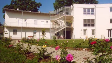 Weißes Mehrfamilienhaus mit Außentreppe und blühendem Rosengarten im Vordergrund.