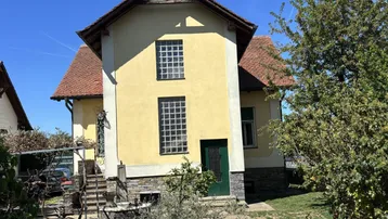 Gelbes Einfamilienhaus mit rotem Ziegeldach und kleinen Fenstern im Grünen.