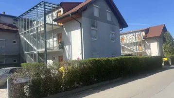 Mehrfamilienhaus mit Außentreppenhaus und gepflegter Hecke im Vordergrund unter blauem Himmel.