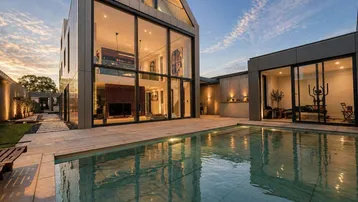 Modernes Haus mit großer Glasfassade, Pool und angrenzendem Fitnessraum bei Sonnenuntergang.