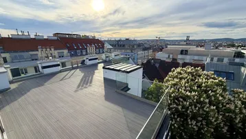 Große Dachterrasse mit modernem Bodenbelag, Glasgeländer und weitem Stadtblick unter sonnigem Himmel.