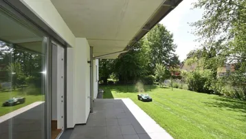 Geräumige Terrasse mit Fliesenboden und Zugang zum gepflegten Garten, wo ein Mähroboter arbeitet.