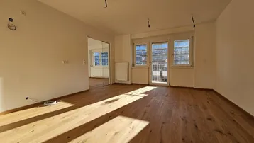 Heller, leerer Wohnraum mit hochwertigem Holzdielenboden, großen Fenstern und Zugang zu einem französischen Balkon.
