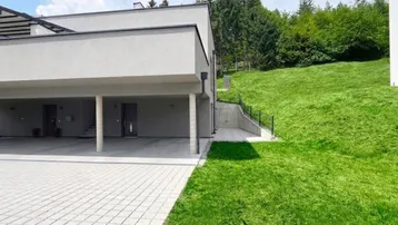 Modernes Gebäude mit Carport und Grünfläche am Hang, umgeben von Bäumen.