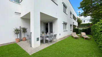 Möblierte Terrasse mit Rasenfläche und Liegestühlen vor dem modernen Gebäude.