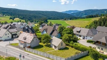 Luftaufnahme eines ländlichen Dorfes mit Häusern, grünen Feldern und Bergen im Hintergrund unter blauem Himmel.