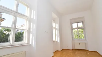 Helles Zimmer mit Parkettboden und großen Fenstern, die viel Tageslicht hereinlassen und einen grünen Ausblick bieten.