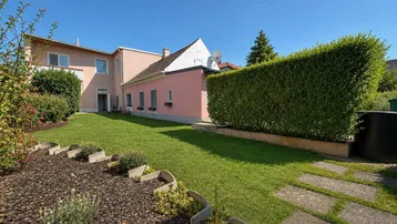 Gepflegter Gartenbereich mit Rasenfläche, Hecken und einem Teil der Hausfassade unter blauem Himmel.