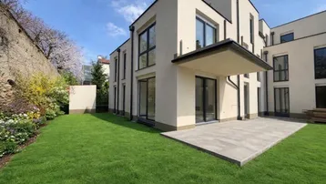 Modernes Gebäude mit heller Fassade, großem Rasenbereich und kleiner Terrasse unter blauem Himmel.