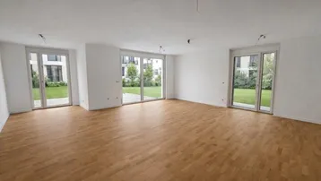 Helles, leeres Zimmer mit Parkettboden und großen Fenstertüren, die Zugang zum Garten bieten.