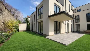 Moderne Wohnanlage mit gepflegtem Rasen, großer Terrasse und angrenzender Steinmauer unter blauem Himmel.