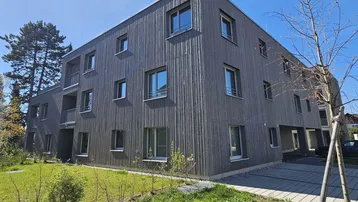 Modernes Mehrfamilienhaus mit Holzfassade und gepflegter Grünfläche unter blauem Himmel.
