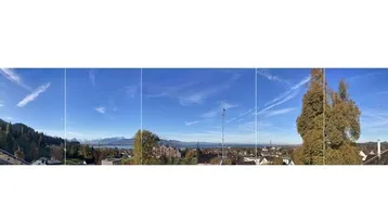 Panoramablick über die Stadt, den See und die umliegenden Berge unter blauem Himmel.