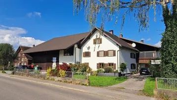 Traditionelles Wohnhaus mit weißer Fassade, braunen Fensterläden und gepflegtem Vorgarten unter blauem Himmel.