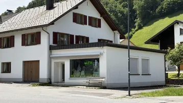 Weißes Gebäude mit roten Fensterläden und einem Ladenlokal im Erdgeschoss, umgeben von grünen Hügeln.