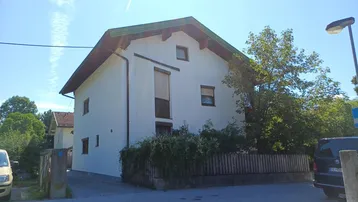 Weißes Wohnhaus mit grünem Satteldach und Holzzaun, umgeben von Bäumen und Sträuchern.