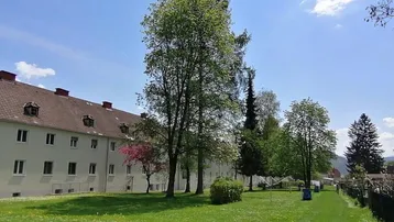 Mehrfamilienhaus mit großer Grünfläche und Bäumen unter blauem Himmel.