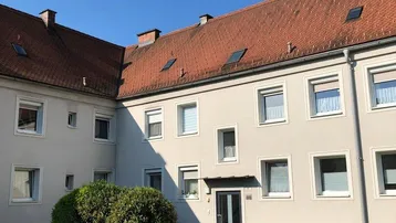 Mehrfamilienhaus mit gepflegter Fassade, roten Ziegeldächern und grünem Vorgarten unter blauem Himmel.