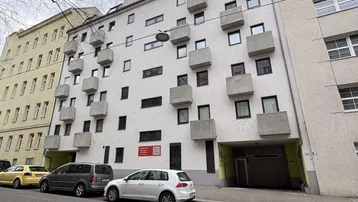 Mehrstöckiges Wohngebäude mit zahlreichen kleinen Balkonen und parkenden Autos auf der Straße.