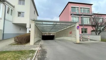 Einfahrt zur Tiefgarage mit Glasüberdachung und angrenzenden Wohngebäuden unter bewölktem Himmel.