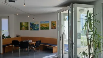 Heller Wartebereich mit bequemer Sitzbank, Tischen und Stühlen, dekoriert mit farbenfrohen Gemälden an der Wand.