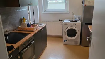Funktionale Küche mit dunklen Schränken, Spüle, Herd und Waschmaschine unter dem Fenster.