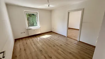 Heller, leerer Raum mit Laminatboden und einem Fenster, das viel Tageslicht hereinlässt.