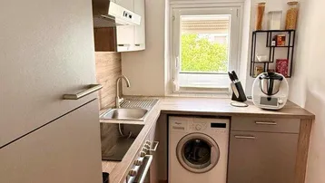 Moderne Küche mit grauen Schränken, Holzarbeitsplatte, integriertem Backofen und Waschmaschine sowie einem Fenster.