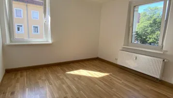 Lichtdurchfluteter Raum mit zwei Fenstern, Parkettboden und Heizkörpern, ideal für Wohn- oder Schlafzimmer.