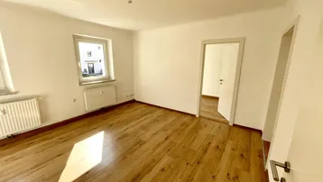 Heller, leerer Raum mit Laminatboden und einem Fenster, das den Blick nach draußen freigibt.