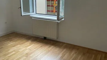 Helles Zimmer mit Parkettboden und einem Fenster, das sich nach außen öffnet.